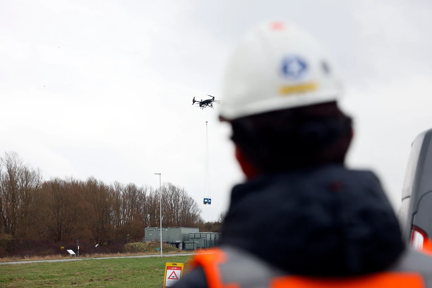 A drone operator wearing a hard hat watches as a drone lifts a cargo payload in an open field, with safety signage and trees in the background under an overcast sky.